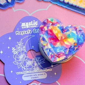 Pu&ograve; includere: Un supporto MagSafe a forma di cuore con un design sfaccettato colorato, appoggiato su una carta viola con il testo "mystic bluu stickers" e l'indirizzo del sito web. La carta ha un design floreale e lunare. Compatibile con telefoni ed e-reader.