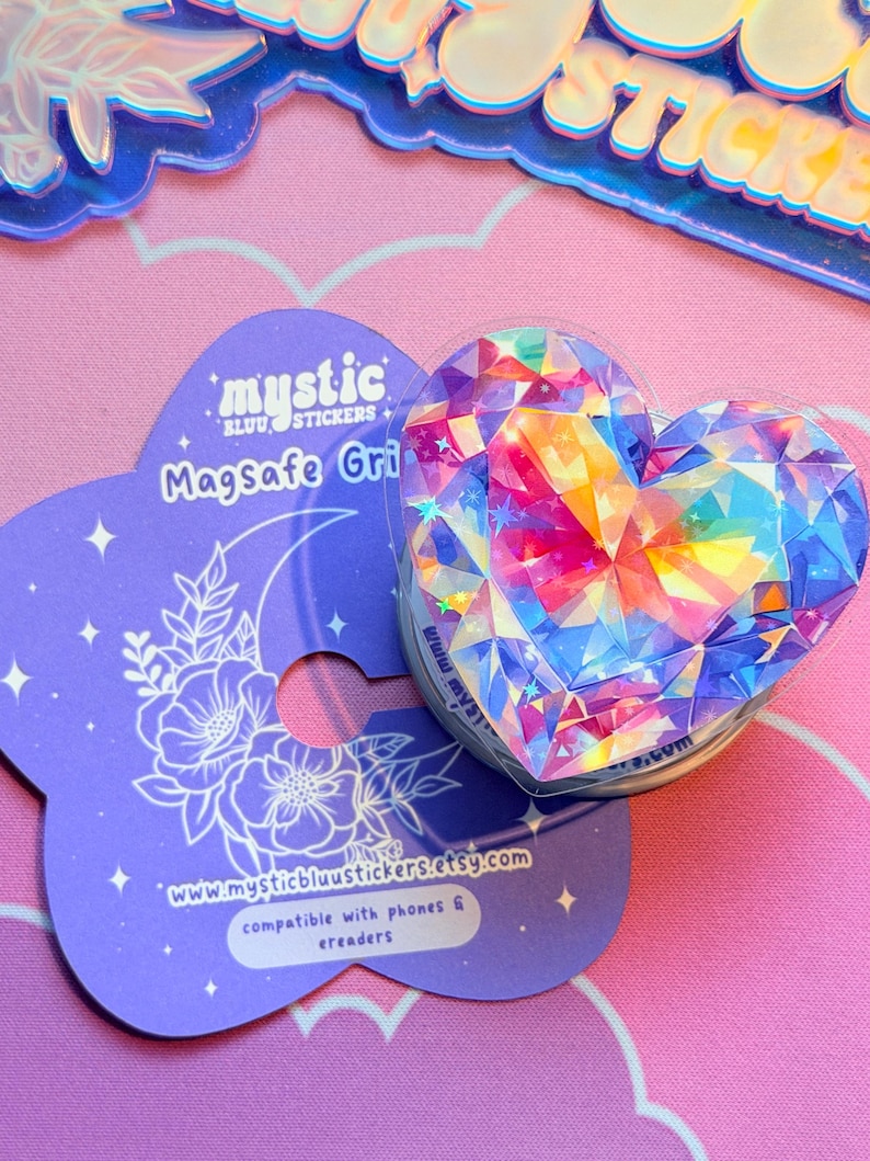Pu&ograve; includere: Un supporto MagSafe a forma di cuore con un design sfaccettato colorato, appoggiato su una carta viola con il testo "mystic bluu stickers" e l'indirizzo del sito web. La carta ha un design floreale e lunare. Compatibile con telefoni ed e-reader.
