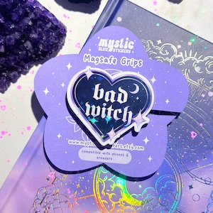 Pode incluir: Uma pega MagSafe roxa em forma de estrela com as palavras "bad witch" num design de coração. A pega está sobre um fundo roxo com o texto "mystic stickers" e "compatible with phones & ereaders".