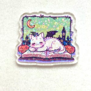 May include: A clear acrylic charm featuring a white cat with bat wings, sleeping on an open book. The background includes a crescent moon, stars, and a spooky castle scene. Two jack-o'-lanterns flank the book, creating a Halloween theme.