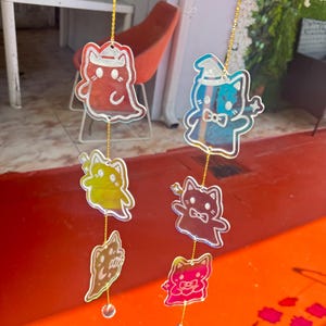 May include: A collection of cat-shaped hanging decorations in red, blue, yellow, and pink. Each ornament is suspended by a gold string and has a clear, reflective surface. The cat designs feature hats, bow ties, and other decorative elements.