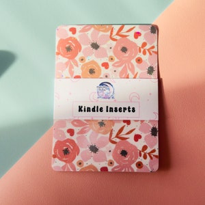 Puede incluir: Un conjunto de insertos para Kindle con estampado floral y el texto "Kindle Inserts" en una etiqueta blanca. Los insertos son de color rosa, naranja y blanco con un acabado brillante.