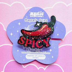 May include: A decorative Magsafe Grip featuring a red chili pepper design with the word "SPICY" in red letters. The grip is on a purple flower-shaped card with the text "mystic stickers". The background is pink.