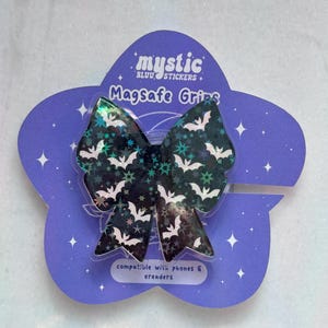 May include: A black bow-shaped MagSafe grip with a pattern of white bats and stars, set against a holographic background. The grip is attached to a purple, flower-shaped backing with the text "mystic" and "MagSafe Grips".