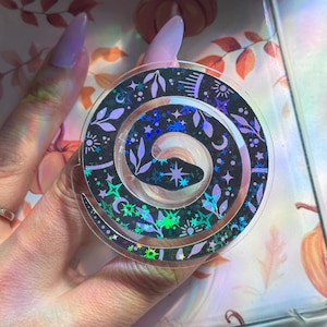 May include: A circular holographic sticker featuring a stylized snake design. The sticker has a black background with a spiral pattern, adorned with stars, leaves, and celestial elements in shades of purple and iridescent colors.