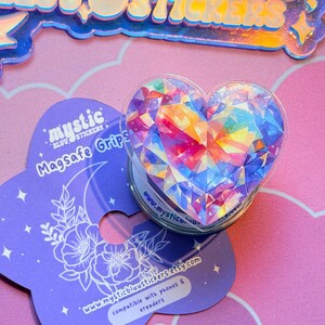Pu&ograve; includere: Un supporto per telefono a forma di cuore con un design a cristallo sfaccettato multicolore. Il supporto &egrave; trasparente con una base bianca, accanto a una carta viola con il testo "mystic blu stickers" e "Magsafe Grips". La carta ha anche un motivo floreale.
