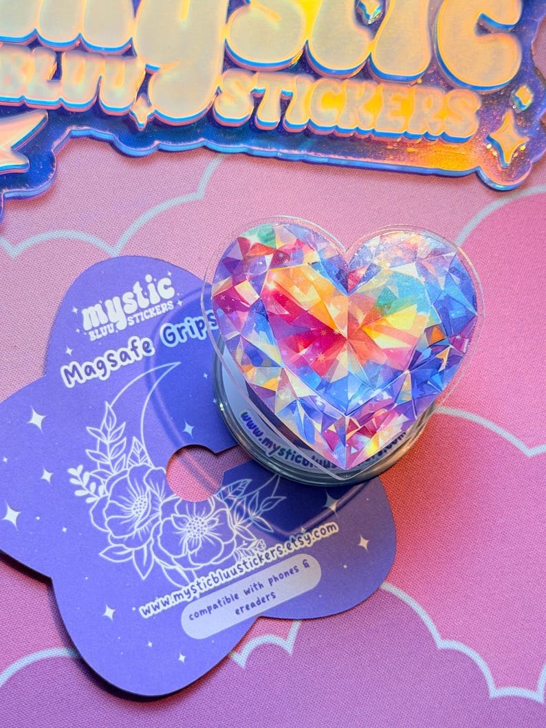 Pu&ograve; includere: Un supporto per telefono a forma di cuore con un design a cristallo sfaccettato multicolore. Il supporto &egrave; trasparente con una base bianca, accanto a una carta viola con il testo "mystic blu stickers" e "Magsafe Grips". La carta ha anche un motivo floreale.