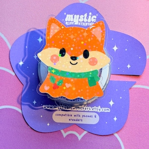 May include: A clear sticker featuring an orange fox with a green scarf and a flower-shaped backing. The sticker has the text "mystic" and "www.mysticbluustickers.etsy.com". The sticker is compatible with phones and e-readers.