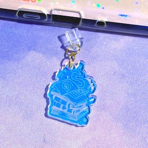 May include: A blue acrylic charm featuring a stack of books with a swirl design on top. The charm is attached to a clear earring post with a gold-colored ring. The background is a light purple fabric.
