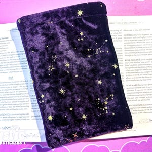 May include: A dark purple velvet pouch with gold star and constellation designs. The pouch is lying on top of an open book with text. The pouch is a drawstring bag, likely for storing small items. The overall theme is mystical and magical.