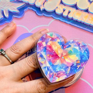 Pu&ograve; includere: Un adesivo a forma di cuore con un design a gemma sfaccettata nei toni del blu, rosa, giallo e viola. L'adesivo &egrave; tenuto in mano. L'adesivo &egrave; compatibile con telefoni ed e-reader.