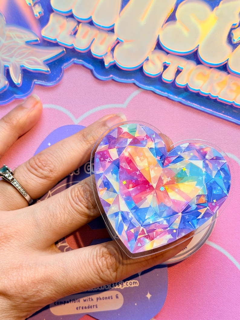 Pu&ograve; includere: Un adesivo a forma di cuore con un design a gemma sfaccettata nei toni del blu, rosa, giallo e viola. L'adesivo &egrave; tenuto in mano. L'adesivo &egrave; compatibile con telefoni ed e-reader.