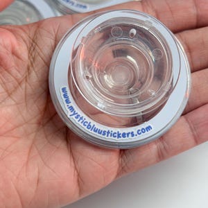 Pu&ograve; includere: Primo piano di adesivi circolari in plastica trasparente con bordo bianco e la scritta "www.mysticbluustickers.com". Gli adesivi hanno un centro trasparente e scanalato.