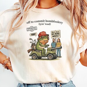 Puede incluir: Camiseta color marfil con la frase "off to commit bombfoolery (lyin' toad)". El gráfico muestra una rana conduciendo un carrito de golf, con palos de golf y un rifle. Dos personas sostienen un cartel que dice "What about NO MORE WARS?"