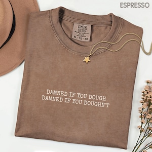 Può includere: T-shirt color espresso con la scritta bianca "DAMNED IF YOU DOUGH DAMNED IF YOU DOUGHN'T". Sono visibili anche una collana con pendente a stella dorata e un cappello marrone. La maglietta è etichettata XL-TG-EG.