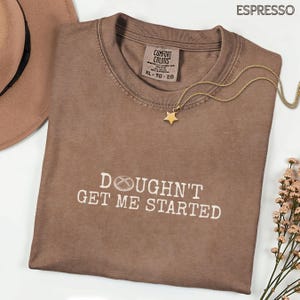 May include: Espresso-coloured t-shirt with the text "Doughn't Get Me Started" in white. A gold star pendant necklace rests on the shirt. A brown hat is partially visible in the upper left corner.
