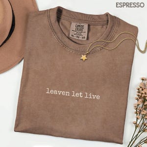 May include: Espresso-coloured t-shirt with the text "leaven let live" in white. A gold star pendant necklace rests on the shirt. A brown hat is partially visible in the upper left corner. The shirt is a Comfort Colors brand, size XL.
