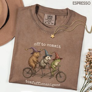 Op de afbeelding: Espresso-kleurig t-shirt met een grillige afbeelding van een capibara, opossum en kikker op een tandemfiets, elk met tovenaars hoeden. De tekst "off to commit tomfufflenanigans" staat eronder. Een gouden sterhangerketting ligt op het shirt.