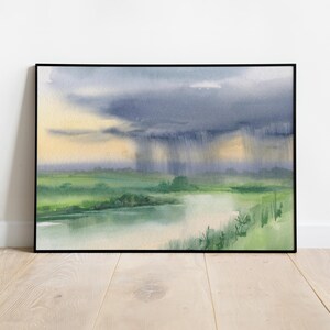May include: Watercolor painting of a landscape with a river and a storm. The sky is a mix of dark blue and yellow, with rain falling over the green fields and water. The artwork is framed in a black frame.