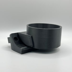May include: Black 3D-printed cup holder with a cylindrical cup section and a rectangular base. The base has a curved section and a hole. The cup holder is designed to hold a cup or other cylindrical object.