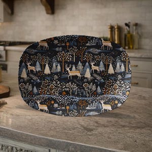 May include: An oval serving platter with a forest scene design. The dark blue platter features a repeating pattern of deer, trees, and foliage in shades of white, gold, and orange. The platter is displayed on a kitchen countertop.