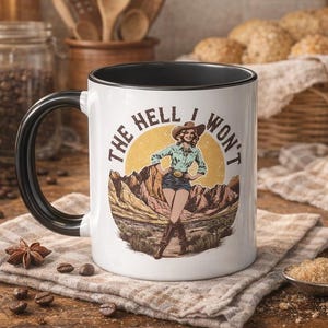 May include: White ceramic mug with a black handle and rim, featuring a retro illustration of a cowgirl with the text "The Hell I Won't." The design includes a cowgirl in a hat, standing in front of a mountain scene. The mug is on a rustic wooden surface.