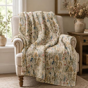 May include: A cream-coloured quilted throw blanket featuring a repeating botanical print of wildflowers in shades of yellow, blue, pink, and green. The blanket is draped over a light-coloured upholstered armchair with visible nailhead trim.