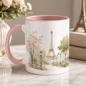 May include: A white ceramic mug with a soft pink interior and handle, featuring a watercolor illustration of the Eiffel Tower in Paris, surrounded by blooming pink cherry blossom trees, green foliage, and black street lamps on a path.