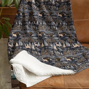 Könnte beinhalten: Eine dunkelblaue Wohndecke mit einem sich wiederholenden Muster aus hellen Hirschen, stilisierten Bäumen und Blattwerk. Die Decke hat ein weiches, weißes, strukturiertes Sherpa-Fleece-Futter, das an der gefalteten Ecke sichtbar ist.