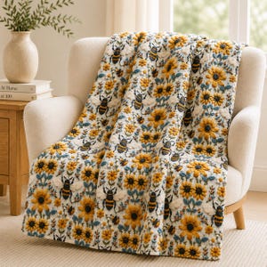 Op de afbeelding: Een witte plaid met een herhalend patroon van gele zonnebloemen en zwart-gele hommels. Het ontwerp bevat kleine blauwe bloemaccenten en groene stengels, wat zorgt voor een dicht, gestructureerd botanisch en insectenmotief.