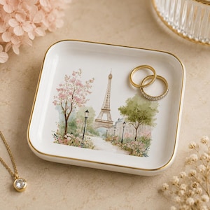May include: A square white ceramic trinket dish with a gold-painted rim, featuring a watercolor-style illustration of the Eiffel Tower surrounded by pink cherry blossoms, green trees, and street lamps, with two gold rings resting on the surface.
