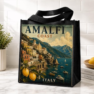 May include: A black tote bag featuring a printed illustration of the Amalfi Coast in Italy. The design displays a coastal town with yellow and orange buildings on a cliffside, blue water with boats, and yellow lemons in the foreground with text.