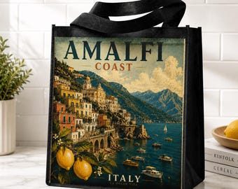 May include: A black tote bag featuring a printed illustration of the Amalfi Coast in Italy. The design displays a coastal town with yellow and orange buildings on a cliffside, blue water with boats, and yellow lemons in the foreground with text.