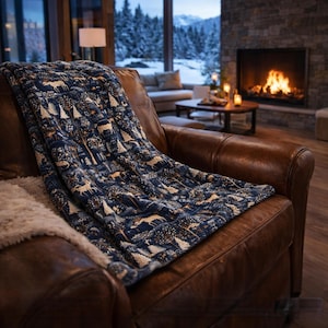 Puede incluir: Una manta azul marino con un estampado de bosque y ciervos sobre un sillón de cuero marrón. La manta presenta un diseño repetido de árboles y ciervos en tonos beige y marrón. La escena está ambientada en un salón acogedor con chimenea.