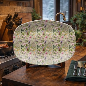 Op de afbeelding: Een ovale serveerschaal met een bloemenpatroon. De schaal heeft een herhalend ontwerp van roze, paarse en gele bloemen met groene bladeren op een lichte achtergrond. De schaal wordt op een houten standaard gepresenteerd.