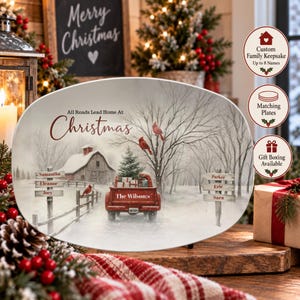 May include: An oval ceramic plate featuring a winter scene with a red pickup truck, a snow-covered barn, and bare trees. The text reads All Roads Lead Home At Christmas and The Wilsons. Two wooden signposts display the names Samantha, Eleanor, Joey, Parker, Eric, and Sara.