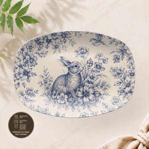 Puede incluir: Una bandeja de servir ovalada con base color crema y un patrón floral azul que presenta un conejo central y un pequeño pájaro. Una etiqueta circular indica que el artículo es apto para lavavajillas, microondas y horno, hecho de polímero.