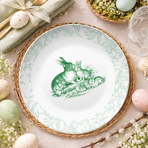 May include: White ceramic plate featuring a green illustration of a rabbit family. The plate has a green floral border and is placed on a woven mat. Easter table setting with eggs and flowers, creating a festive atmosphere.