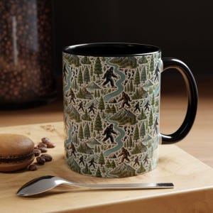 Puede incluir: Una taza de café con asa negra y un diseño temático de Bigfoot. El diseño presenta figuras de Bigfoot, árboles, montañas y un río sobre un fondo crema. La taza tiene un interior negro y está sobre una superficie de madera.