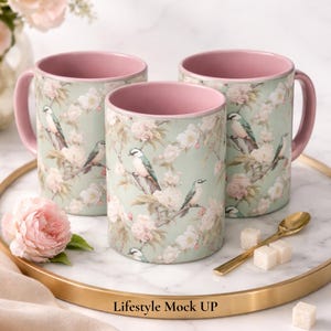 May include: Three ceramic mugs with a light green exterior featuring a pattern of birds and pale pink flowers. The interior and handle of each mug are a solid dusty pink color. The text "Lifestyle Mock UP" is printed at the bottom of the image.