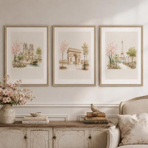 May include: A set of three framed wall art prints featuring watercolor illustrations of Paris landmarks including Notre Dame, the Arc de Triomphe, and the Eiffel Tower, each surrounded by blooming pink cherry blossom trees and greenery.