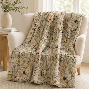 May include: A cream-colored quilted throw blanket featuring an all-over embroidered pattern of wildflowers, including white daisies, yellow blossoms, and various green foliage, draped over a light-colored upholstered armchair.