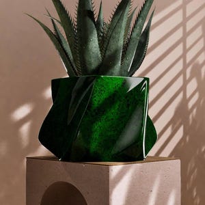 May include: A dark green, geometric-shaped planter holding a spiky succulent plant. The glossy planter sits on a light-colored, square pedestal with an arched cutout. The background is a neutral color with shadows.