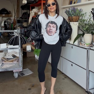 May include: A person wearing a white graphic t-shirt with a portrait of a man smoking a cigarette, black leggings, a black leather jacket, and dark sunglasses. They are also wearing dark brown pointed-toe heels.