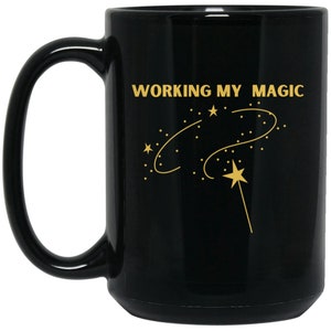 May include: Black ceramic mug with a gold design. The mug has a white handle and the text "WORKING MY MAGIC" with a gold star and a wand.