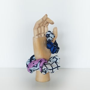 May include: A wooden hand mannequin displays several patterned scrunchies. The scrunchies feature various designs, including polka dots, geometric shapes, and floral patterns. The fabric colors include blue, pink, and white.