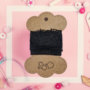 Può includere: Una bobina di nastro di pizzo nero su una bobina decorativa di cartone a forma di nuvola. La bobina ha un piccolo foro nella parte superiore e un logo nella parte inferiore. Lo sfondo è rosa con elementi decorativi.