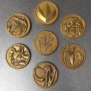 Può includere: Una collezione di sette monete color oro, ognuna con un design in rilievo unico. Le monete presentano vari animali preistorici e simboli, tra cui un Tyrannosaurus Rex, una tigre dai denti a sciabola e un Triceratopo. Le monete sono disposte su una superficie grigia.