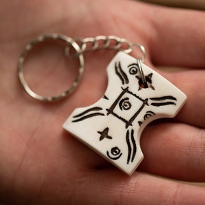 May include: A white, hammer-shaped keychain pendant with black etched designs, including swirls, lines, and a central square. The pendant is attached to a silver chain and keyring. The background is a blurred hand.