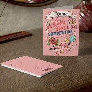Puede incluir: Una tarjeta de felicitación rosa con el texto "Cute But Competitive" y varias ilustraciones, incluyendo cerezas, una rosa, gafas de sol y una corona. La tarjeta está abierta, revelando un interior en blanco, y está colocada sobre una superficie de madera.